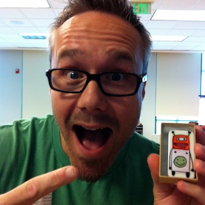 Puede incluir: Un hombre con una camisa verde y barba de chivo sonríe y sostiene una pequeña caja con una ilustración de robot. El robot es naranja, rojo y blanco con una cara amarilla y ojos negros. La caja es blanca con bordes negros.