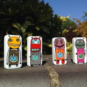 May include: Four ceramic figurines of cartoon characters with different colored bodies and faces. Each figurine has a large, round button on its chest. The figurines are standing on a gray surface.