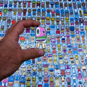 May include: A hand holds a small, colorful, cartoon-like character with a pink face, green hair, and a white body. The character is surrounded by a background of hundreds of similar characters in various colors and designs.