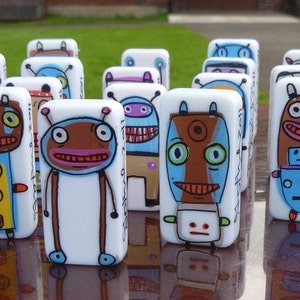 Puede incluir: Una colección de dominós pintados a mano con personajes de robots coloridos y caprichosos. Cada dominó es blanco con contornos negros y presenta diseños de robots únicos con diferentes colores, patrones y expresiones.