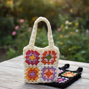 May include: A cream-colored crochet tote bag featuring four square granny motifs in shades of orange, yellow, pink, and purple. A second, similar black crochet bag with colorful granny squares is positioned flat underneath the upright cream bag.