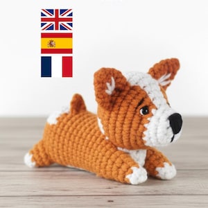 May include: A crocheted corgi dog in shades of orange, white, and black, lying down on a wooden surface. The dog has a detailed design with visible stitches. The text "Crochet PDF Pattern" is at the top of the image, with flags of the UK, Spain, and France.