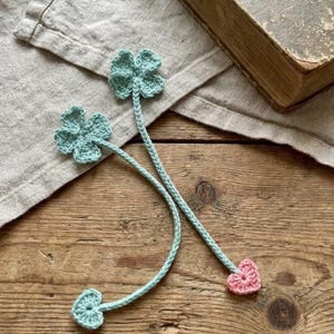 Op de afbeelding: Twee handgemaakte gehaakte bladwijzers. Elke bladwijzer heeft een lichtblauwe klaver met vier bladeren, een gevlochten steel en een roze hart. Ze liggen op een houten oppervlak naast een vintage boek.