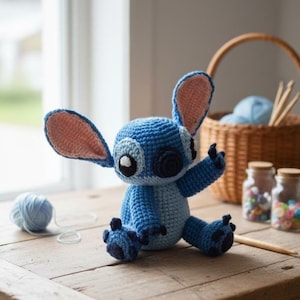 Può includere: Bambola Stitch blu all'uncinetto fatta a mano con grandi orecchie e un'espressione amichevole. La bambola è seduta su una superficie di legno, con una palla di filo blu chiaro e un cestino sullo sfondo. Le orecchie della bambola sono rosa all'interno.