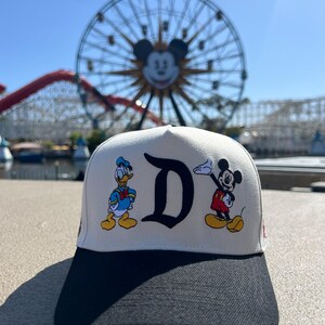 Op de afbeelding: Een wit en zwart baseball cap met Donald Duck en Mickey Mouse. De pet heeft een grote zwarte "D" en een zwarte klep. Op de achtergrond een reuzenrad en een achtbaan.