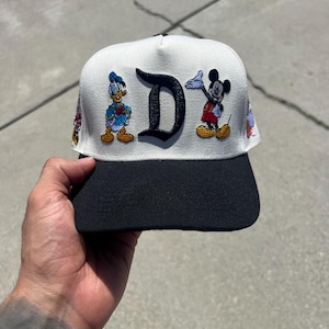 May include: A baseball cap with a white crown and a black brim. The front features an embroidered black letter D flanked by a colourful Donald Duck figure on the left and a Mickey Mouse figure on the right. Additional small character patches are visible.