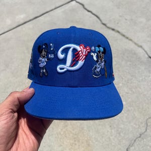 May include: A royal blue baseball cap featuring a large white embroidered letter D with a red baseball graphic. The cap is decorated with two embroidered Mickey Mouse figures, one holding a foam finger and the other holding a baseball bat.