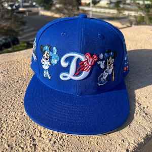 May include: A royal blue baseball cap featuring an embroidered white letter D with a red Mickey Mouse icon baseball design. The cap includes embroidered figures of Mickey Mouse and Minnie Mouse wearing blue sports gear on the front panels.