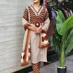 May include: A two-piece brown and white patterned outfit with a matching scarf. The top has a V-neck and a decorative circular accent at the waist. The pants are a matching print. The outfit is worn with black sandals.