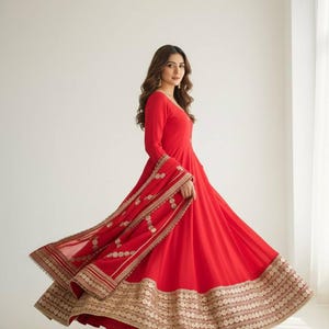 May include: A flowing, red dress with long sleeves and a full skirt. The dress features intricate gold embroidery along the hem and sleeves. The model is wearing the dress in a studio setting with natural light.