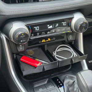 Könnte beinhalten: Schwarzer Kunststoff-Autokonsolen-Organizer mit zwei Fächern. Ein Fach enthält ein rotes Feuerzeug, einen Stift und eine Münze. Das andere enthält ein silbernes und graues Kabel. Die Klimaanzeige des Autos zeigt 21 Grad Celsius an.