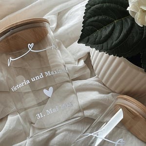 May include: Two clear glass jars with wooden lids, personalized with white text and a heart design. One jar reads "Victoria und Maxim" and "31. Mai 2025." The other reads "02. August 2025" and "Alles Liebe zur Hochzeit."