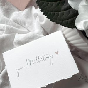 May include: A white card with the words "zum Muttertag" in elegant script, and a small pink heart. The card is set against white fabric, a pink envelope, and dark green leaves, creating a soft, natural aesthetic.