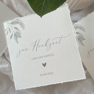 Op de afbeelding: Vierkante witte huwelijksuitnodiging met de woorden "zur Hochzeit" en de namen "Lina und Marcel" in grijs gedrukt. Een hartsymbool en de datum 30.08.2025 staan ook op de uitnodiging. De uitnodiging heeft een bloemenontwerp.