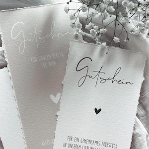 Può includere: Primo piano di tre buoni regalo di carta strutturata. Il buono superiore recita "Gutschein" in corsivo. Il secondo buono ha il testo "FÜR EIN GEMEINSAMES FRÜHSTÜCK" e "HAPPY BIRTHDAY, MAMAT". I buoni sono su un tessuto bianco.