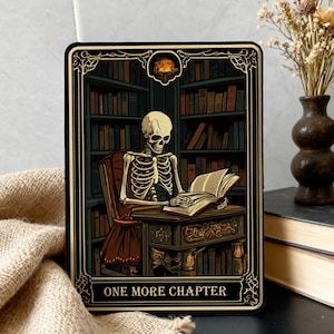 Può includere: Carta rettangolare con uno scheletro che legge in una biblioteca. Bordo nero con dettagli dorati e la scritta "ONE MORE CHAPTER". Lo scheletro è seduto a una scrivania di legno.