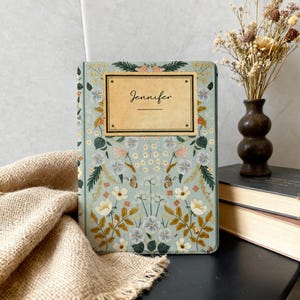 May include: A personalized journal with a floral design. The cover is light blue with a gold-colored rectangle that reads "Jennifer". The floral pattern includes white, pink, and yellow flowers, with green leaves and butterflies.