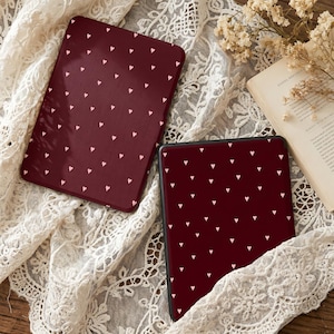 Puede incluir: Dos fundas de lector de libros electrónicos de color burdeos con un patrón de pequeños corazones rosa claro. Las fundas son rectangulares y tienen una superficie lisa. Una funda está abierta, revelando la pantalla. Las fundas están sobre una superficie de encaje blanco.