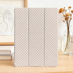 Peut inclure: Un étui de tablette crème avec un motif répété de petits points rouges espacés uniformément. L'étui est plié ouvert. L'arrière-plan comprend un vase de fleurs séchées et un dessin encadré.