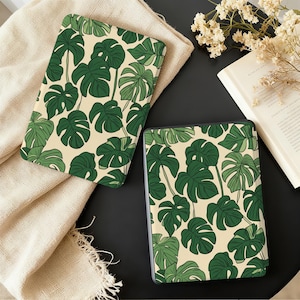 Op de afbeelding: Twee rechthoekige hoesjes met een crèmekleurige achtergrond en een herhalend patroon van grote, groene monstera bladeren. Eén hoesje is open en onthult een zwart interieur. Het andere hoesje is gesloten. De hoesjes liggen op een zwart oppervlak.