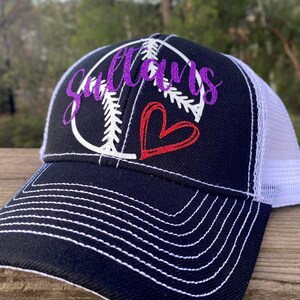 May include: A black and white baseball themed trucker hat with a purple glitter design that says "Sultans" and a red heart. The hat has a white mesh back.