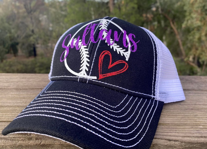 May include: A black and white baseball themed trucker hat with a purple glitter design that says "Sultans" and a red heart. The hat has a white mesh back.