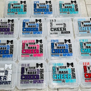 May include: Twelve clear plastic containers with lids, each decorated with a different cheerleading design. The designs feature a variety of colors, including blue, red, purple, pink, and teal. Each container has a name at the top, such as "Sienna", "Evie", "Lily", and "Abby".