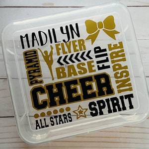 May include: Clear plastic storage box with a gold and black design. The design includes the words "Madilyn", "Cheer", "All Stars", "Pyramid", "Flyer", "Base", "Flip", "Inspire", and "Spirit". There is also a gold bow and a gold star.