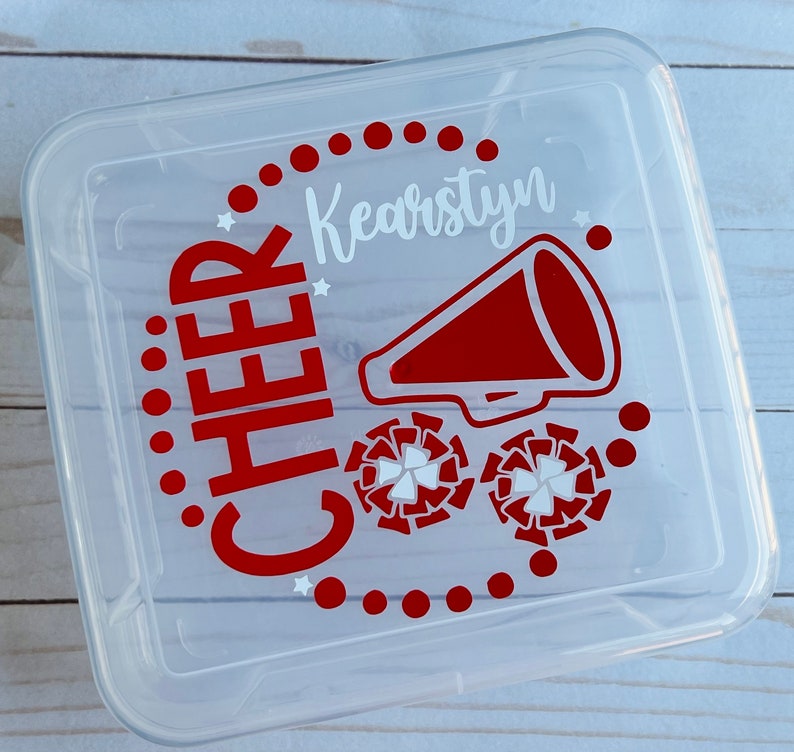 Puede incluir: Contenedor de almacenamiento de pl&aacute;stico transparente con un dise&ntilde;o rojo y blanco. El dise&ntilde;o incluye las palabras "CHEER" y "Kearstyn" junto con un meg&aacute;fono y pompones de animadora.