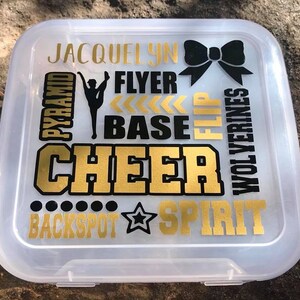 May include: A clear plastic storage box with a gold and black design. The design includes the words "JACQUELYN", "PYRAMID", "FLYER", "BASE", "FLIP", "CHEER", "WOLVERINES", "BACKSPOT", and "SPIRIT". There is also a black bow and a gold star.