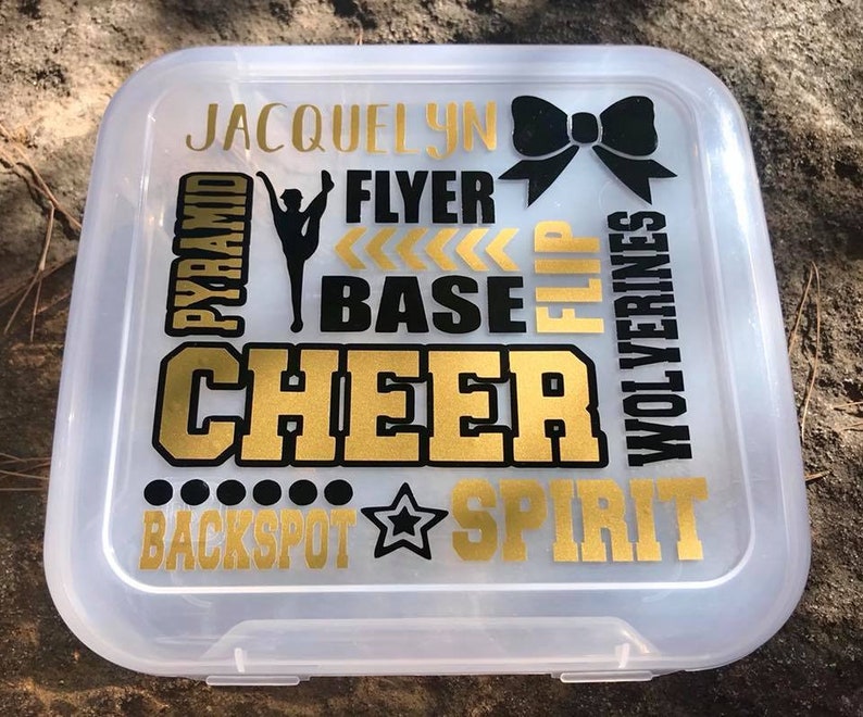 May include: A clear plastic storage box with a gold and black design. The design includes the words "JACQUELYN", "PYRAMID", "FLYER", "BASE", "FLIP", "CHEER", "WOLVERINES", "BACKSPOT", and "SPIRIT". There is also a black bow and a gold star.