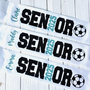 Op de afbeelding: Drie witte sjerpen met zwarte glittertekst die "Senior 2025" zegt en een turquoise glitter "2025". Elke sjerp heeft een andere naam: Claire, Molly en Emma. Elke sjerp heeft ook een zwart-witte voetbalafbeelding.