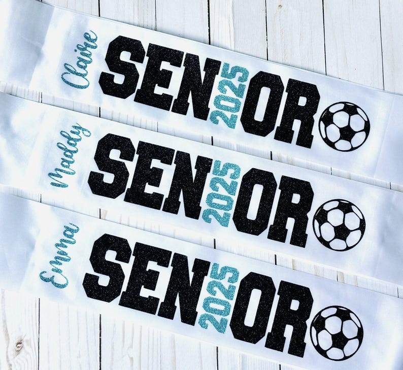 Op de afbeelding: Drie witte sjerpen met zwarte glittertekst die "Senior 2025" zegt en een turquoise glitter "2025". Elke sjerp heeft een andere naam: Claire, Molly en Emma. Elke sjerp heeft ook een zwart-witte voetbalafbeelding.