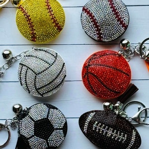 May include: A set of six keychains featuring different sports balls, each covered in sparkling rhinestones. The balls include a baseball, basketball, football, soccer ball, volleyball, and softball.