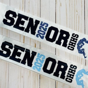 May include: Two white satin sashes with the text "Senior 2025 CHHS" in black lettering. The sashes have a blue glitter design and a wrestling silhouette.