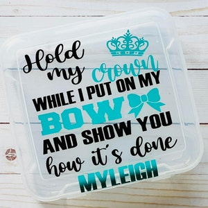 May include: A clear plastic storage box with a turquoise crown, bow, and text that reads "Hold my crown while I put on my bow and show you how it's done MYLEIGH".