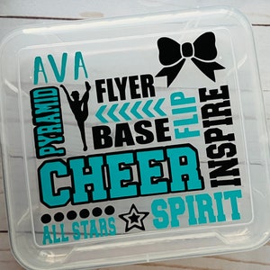 May include: A clear plastic storage box with a teal and black design. The design includes the words "cheer", "pyramid", "flyer", "base", "flip", "inspire", "spirit", and "all stars". The name "Ava" is also included in the design.