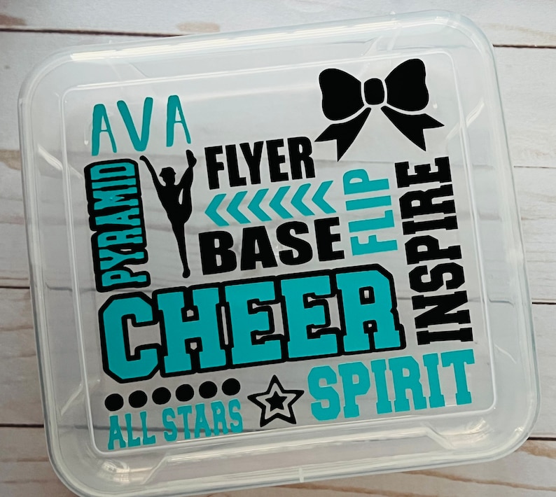 May include: A clear plastic storage box with a teal and black design. The design includes the words "cheer", "pyramid", "flyer", "base", "flip", "inspire", "spirit", and "all stars". The name "Ava" is also included in the design.