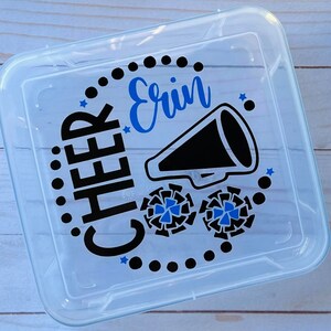 Puede incluir: Un contenedor de almacenamiento de pl&aacute;stico transparente con un dise&ntilde;o negro y azul. El dise&ntilde;o incluye las palabras "Cheer Erin" y un meg&aacute;fono con pompones de animadora.