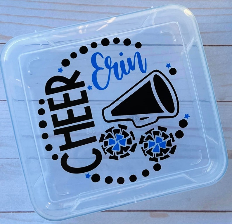 Puede incluir: Un contenedor de almacenamiento de pl&aacute;stico transparente con un dise&ntilde;o negro y azul. El dise&ntilde;o incluye las palabras "Cheer Erin" y un meg&aacute;fono con pompones de animadora.