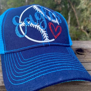 May include: A blue and turquoise trucker hat with a white baseball graphic and the word "Eagles" in glitter. A red heart is also on the hat.