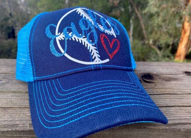 May include: A blue and turquoise trucker hat with a white baseball graphic and the word "Eagles" in glitter. A red heart is also on the hat.