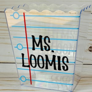 Puede incluir: Un recipiente de pl&aacute;stico transparente con un borde festoneado y un dise&ntilde;o de l&iacute;neas azules y blancas. El recipiente tiene el texto "MS. LOOMIS" escrito en negro.