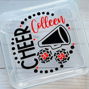 Puede incluir: Contenedor de almacenamiento de pl&aacute;stico transparente con un dise&ntilde;o en negro y rojo. El dise&ntilde;o incluye las palabras "CHEER Colleen" y un meg&aacute;fono negro con estrellas rojas. Tambi&eacute;n hay dos pompones rojos y negros.