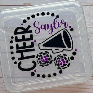 Puede incluir: Contenedor de almacenamiento de pl&aacute;stico transparente con un dise&ntilde;o negro y morado. El dise&ntilde;o incluye las palabras "CHEER" y "Saylor" junto con un meg&aacute;fono y pompones de animadora.