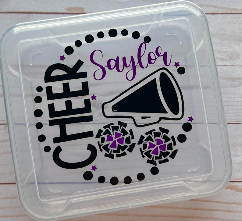 Puede incluir: Contenedor de almacenamiento de pl&aacute;stico transparente con un dise&ntilde;o negro y morado. El dise&ntilde;o incluye las palabras "CHEER" y "Saylor" junto con un meg&aacute;fono y pompones de animadora.