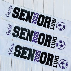 Op de afbeelding: Drie witte afstudeersjaals met zwarte tekst die "Senior 2025" en de naam van elke afgestudeerde leest. Elke sjaal heeft een paarse glittervoetbal.
