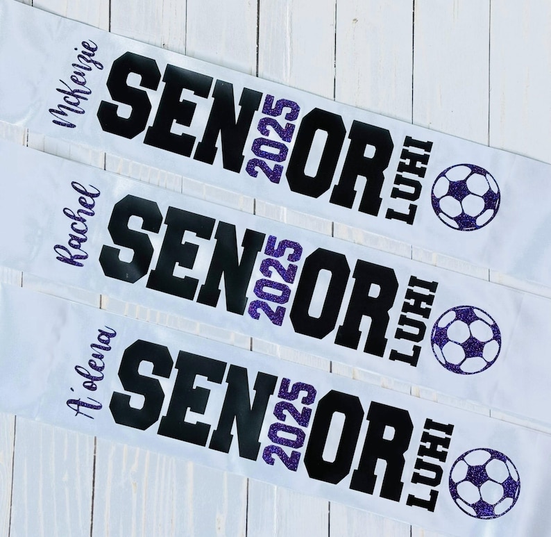 Op de afbeelding: Drie witte afstudeersjaals met zwarte tekst die "Senior 2025" en de naam van elke afgestudeerde leest. Elke sjaal heeft een paarse glittervoetbal.