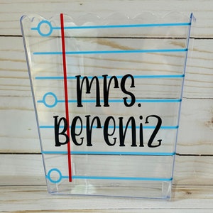 Puede incluir: Contenedor de pl&aacute;stico transparente con l&iacute;neas azules y una l&iacute;nea roja a un lado. El contenedor tiene una parte superior festoneada y el texto "mrs. bereniz" est&aacute; impreso en negro.