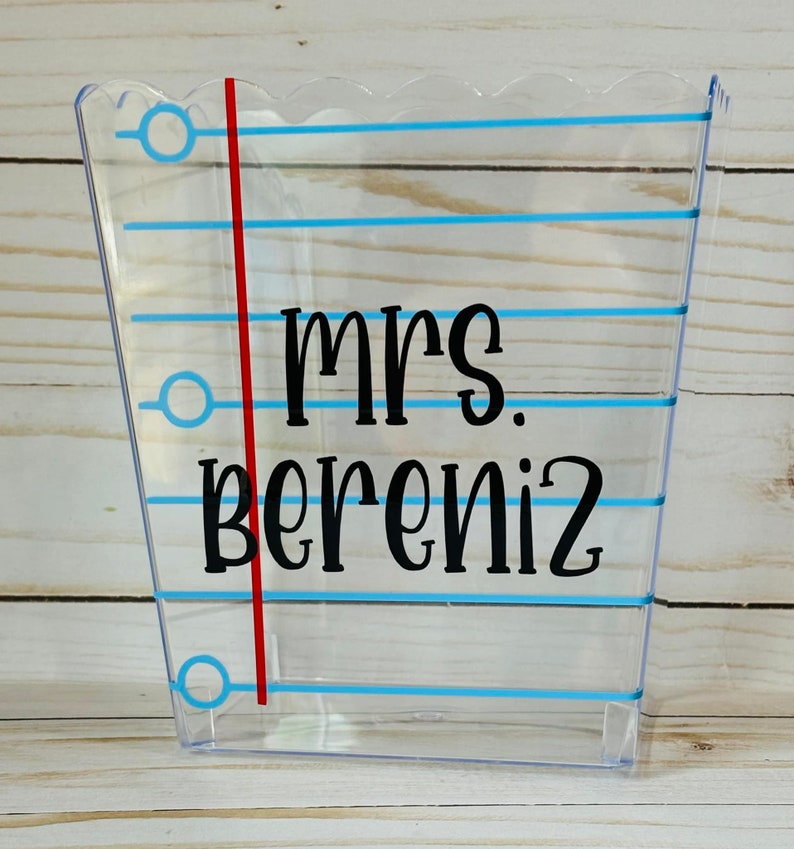 Puede incluir: Contenedor de pl&aacute;stico transparente con l&iacute;neas azules y una l&iacute;nea roja a un lado. El contenedor tiene una parte superior festoneada y el texto "mrs. bereniz" est&aacute; impreso en negro.